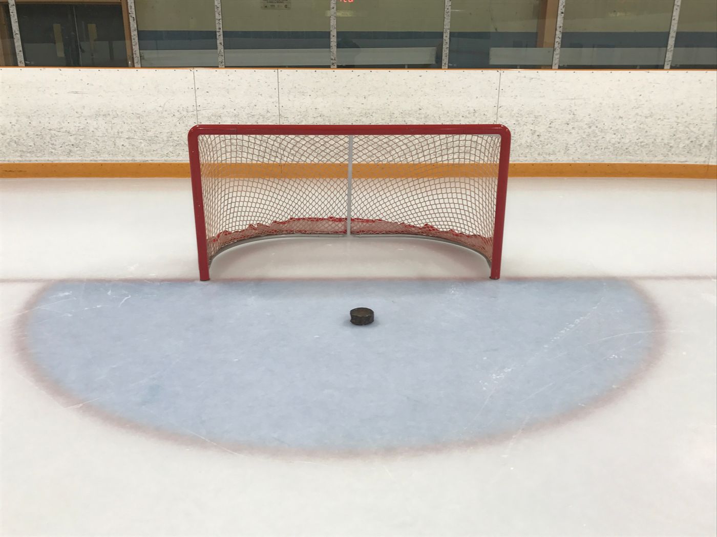 A Blind Hockey net on the goal line, seen from the front: red posts and a crossbar set lower than a standard net, with the large Blind Hockey puck sitting in the blue goal crease in front of it.