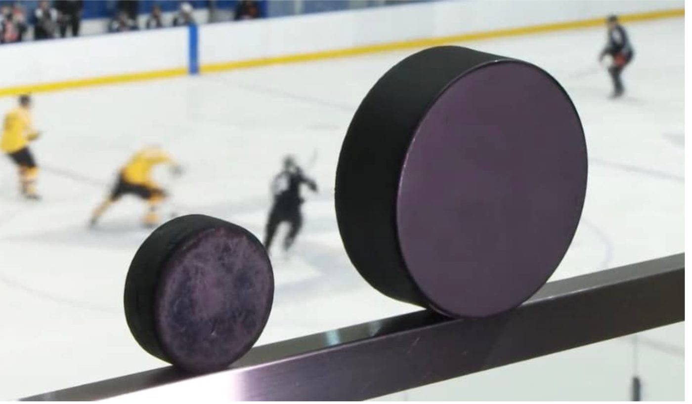 A standard hockey puck and a Blind Hockey puck standing side by side on the rink boards. The Blind Hockey puck on the right is nearly twice as wide and noticeably thicker. A game is being played on the ice behind them.