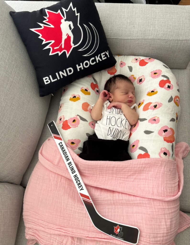 Bianca is sleeping in her chair and there’s a black pillow next to her with the Canadian Blind Hockey logo and a mini stick at her feet