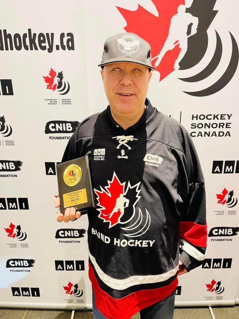 Mario Ros holding his award during the ceremony stands infront of the Canadian Blind Hockey backdrop